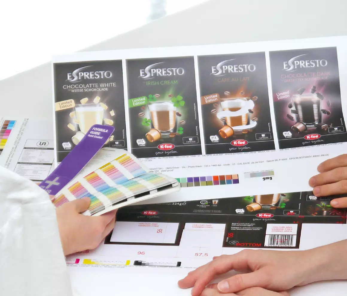 The hands of two women. one holds a Pantone color fan in her hand and compares the colors with the design proof on the table.