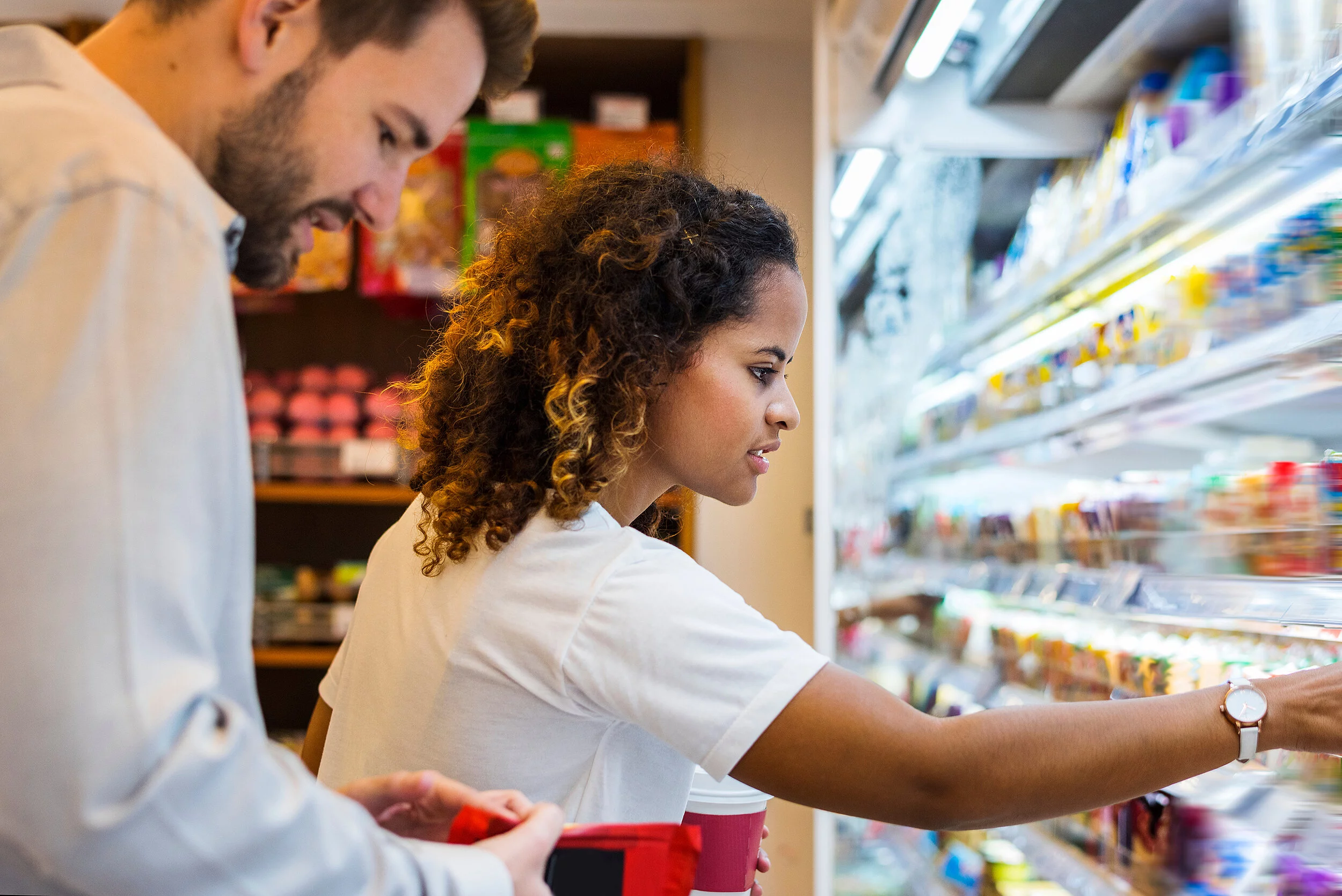 A man and a woman in front of the refrigerated shelf. She reaches for a package, he scrolls on his mobile phone.