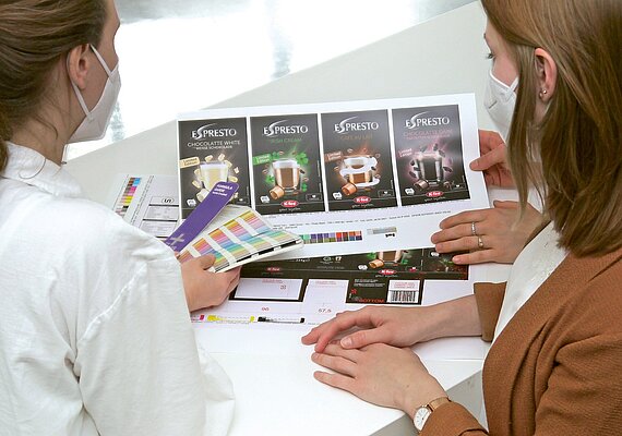 The hands of two women. one holds a Pantone color fan in her hand and compares the colors with the design proof on the table.